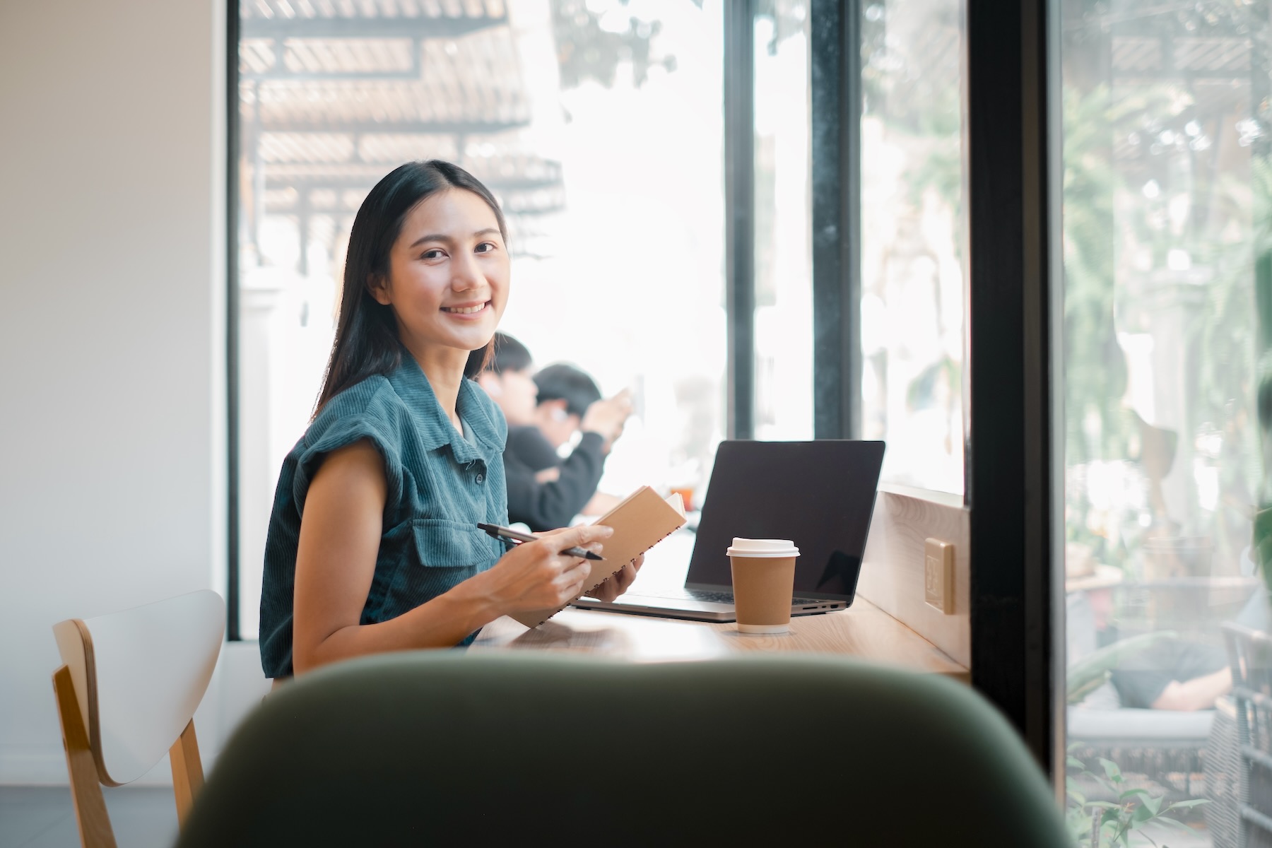 A woman is sitting at a table with a laptop and a notebook. She is smiling and she is enjoying her time. The scene suggests a relaxed and comfortable atmosphere. The woman might be working